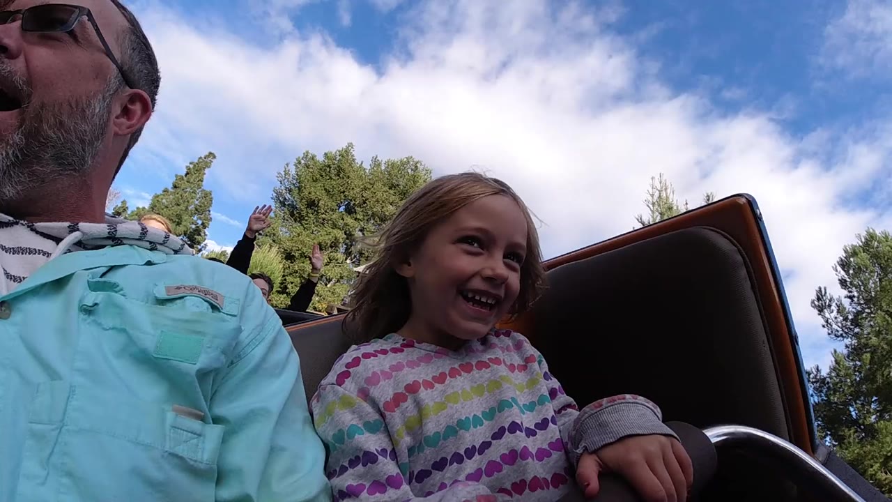 At Disneyland California, a bearded man in sunglasses is cropped at left while a young girl laughs beside a dark coaster seat under blue sky and clouds.