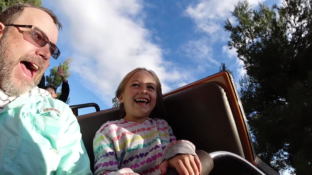 At Disneyland California, a father in sunglasses and a smiling young girl sit close to the camera in a roller coaster car with trees and clouds behind them.