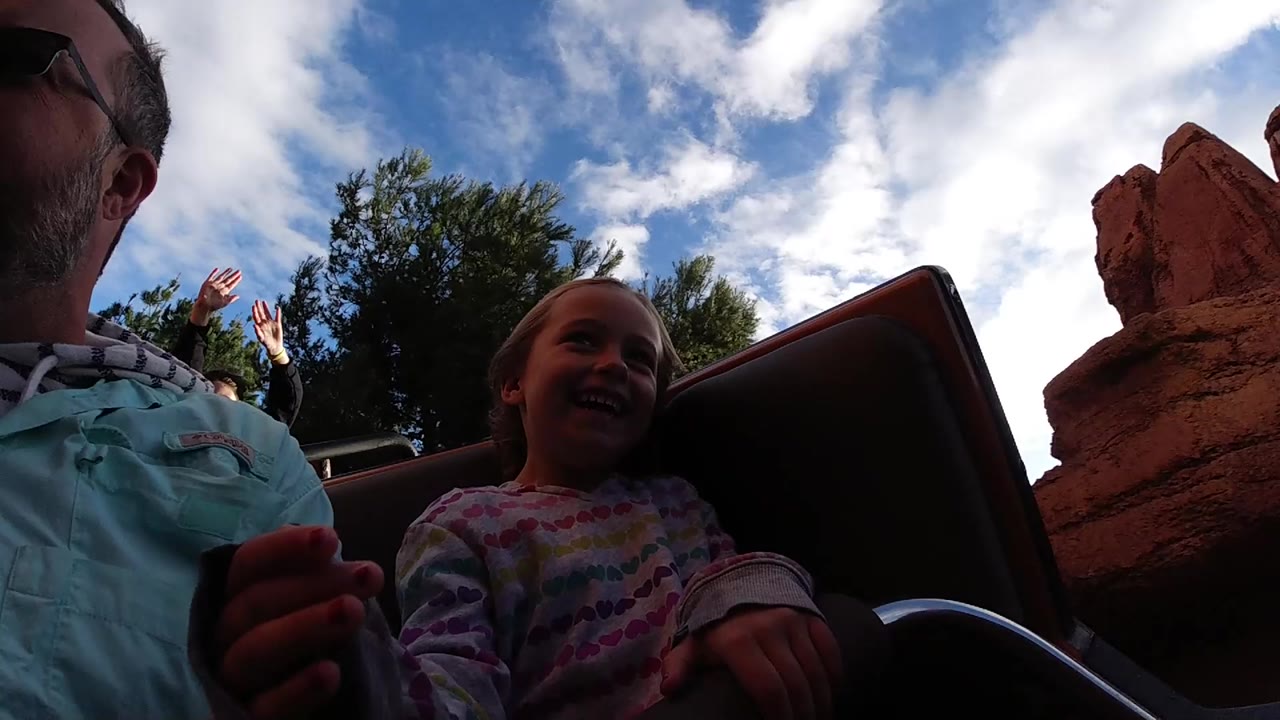 At Disneyland California, a young girl smiles beside an adult man in a ride vehicle, with red rockwork, trees, and bright clouds behind them.