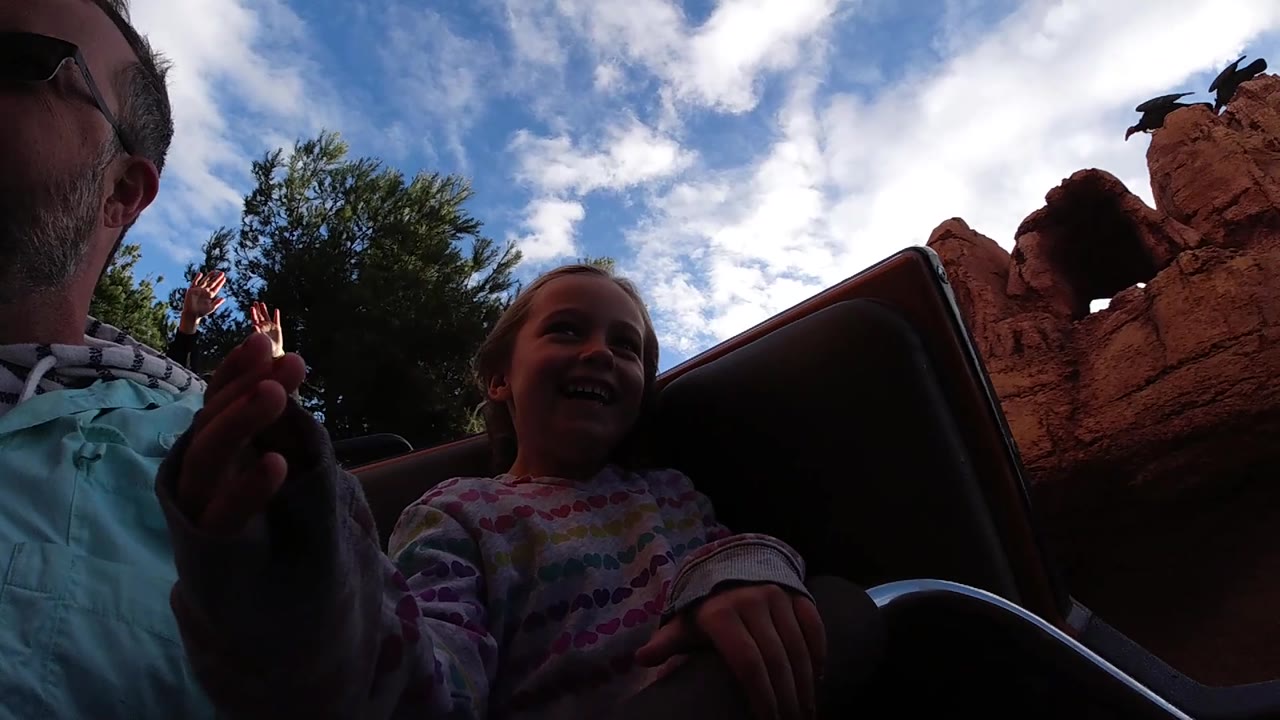 At 0:02, the girl laughs in the ride vehicle while a hand lifts near the lens and red themed rocks and clouded sky fill the background.