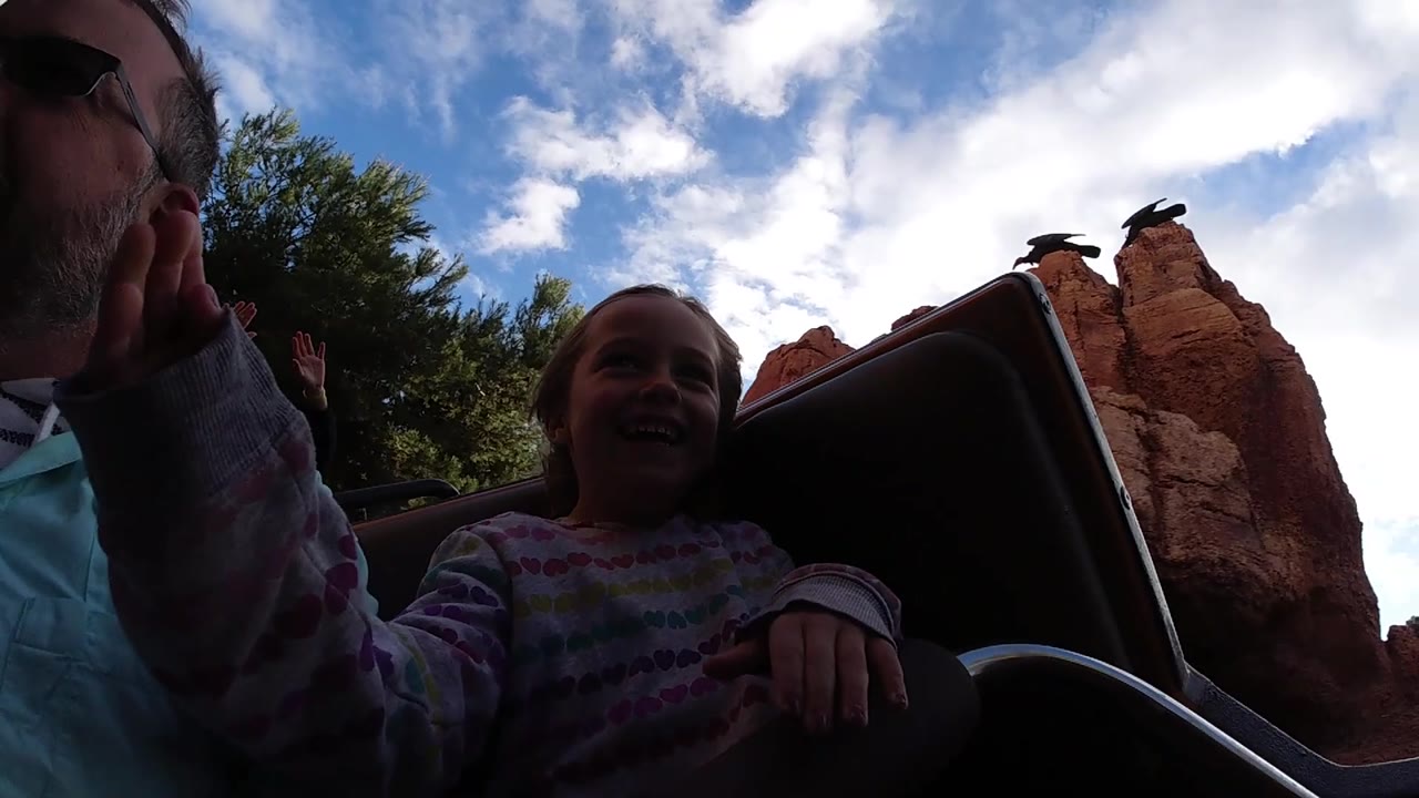 Adult man and girl seated on ride with hands raised near red rock formations.