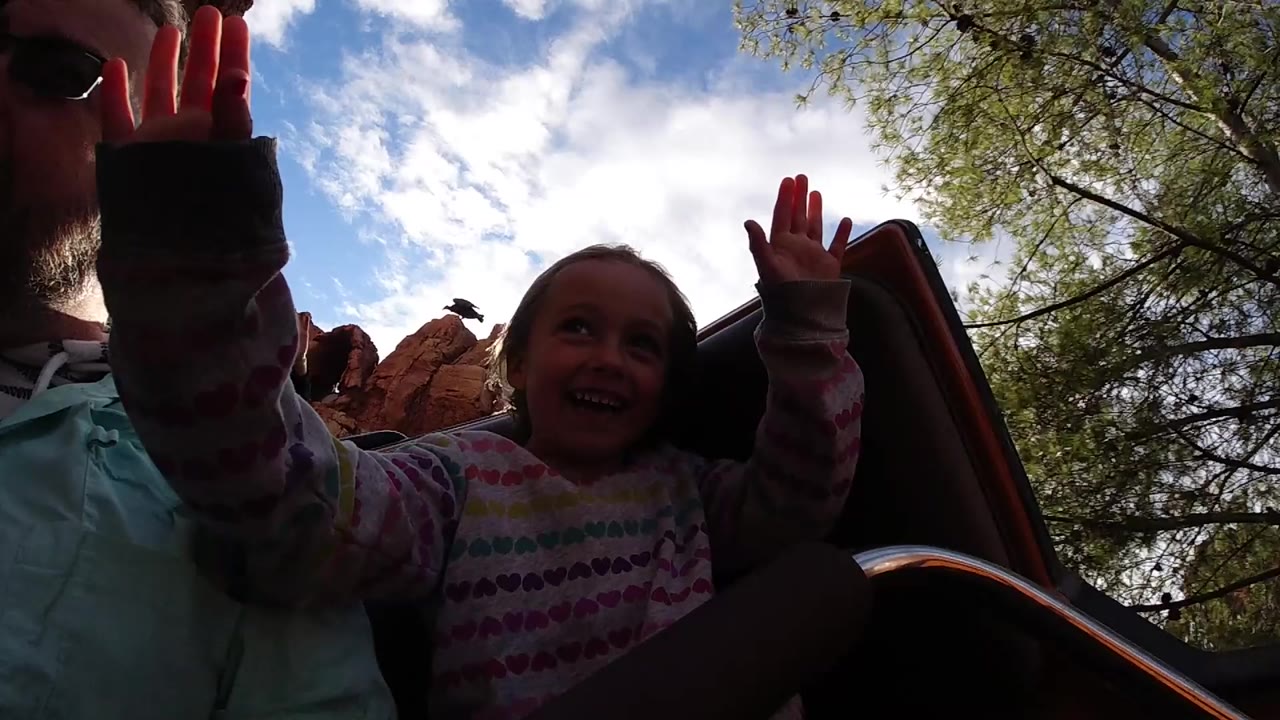 The young girl smiles while raising one hand beside an adult rider, with the roller coaster seat edge, trees, clouds, and red rock scenery visible around them at Disneyland California.