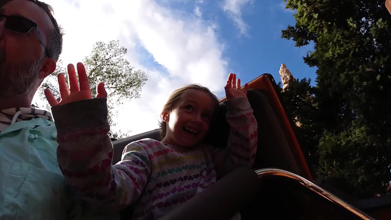 At Disneyland California, a man in sunglasses and a young girl sit close to a low onboard camera with raised hands, trees and bright sky behind them.