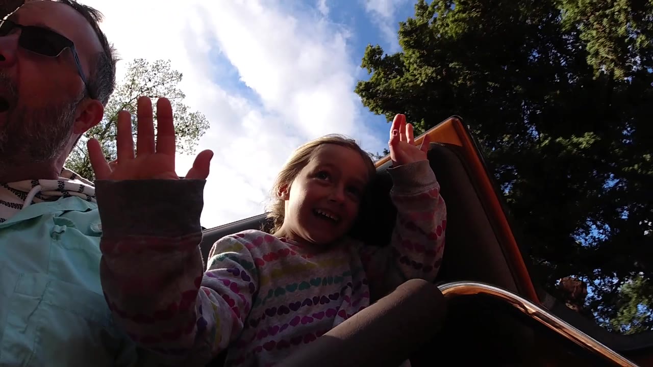 The man and young girl remain close to the low camera as raised hands fill the foreground, with tree canopy and sky above the Disneyland California ride.