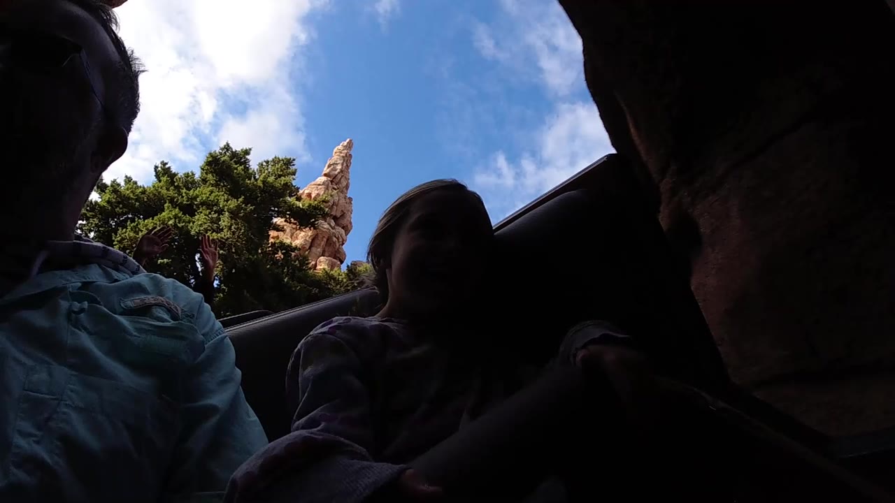 At Disneyland California, silhouetted riders fill the foreground as trees and a narrow tan rock spire pass behind the coaster car.