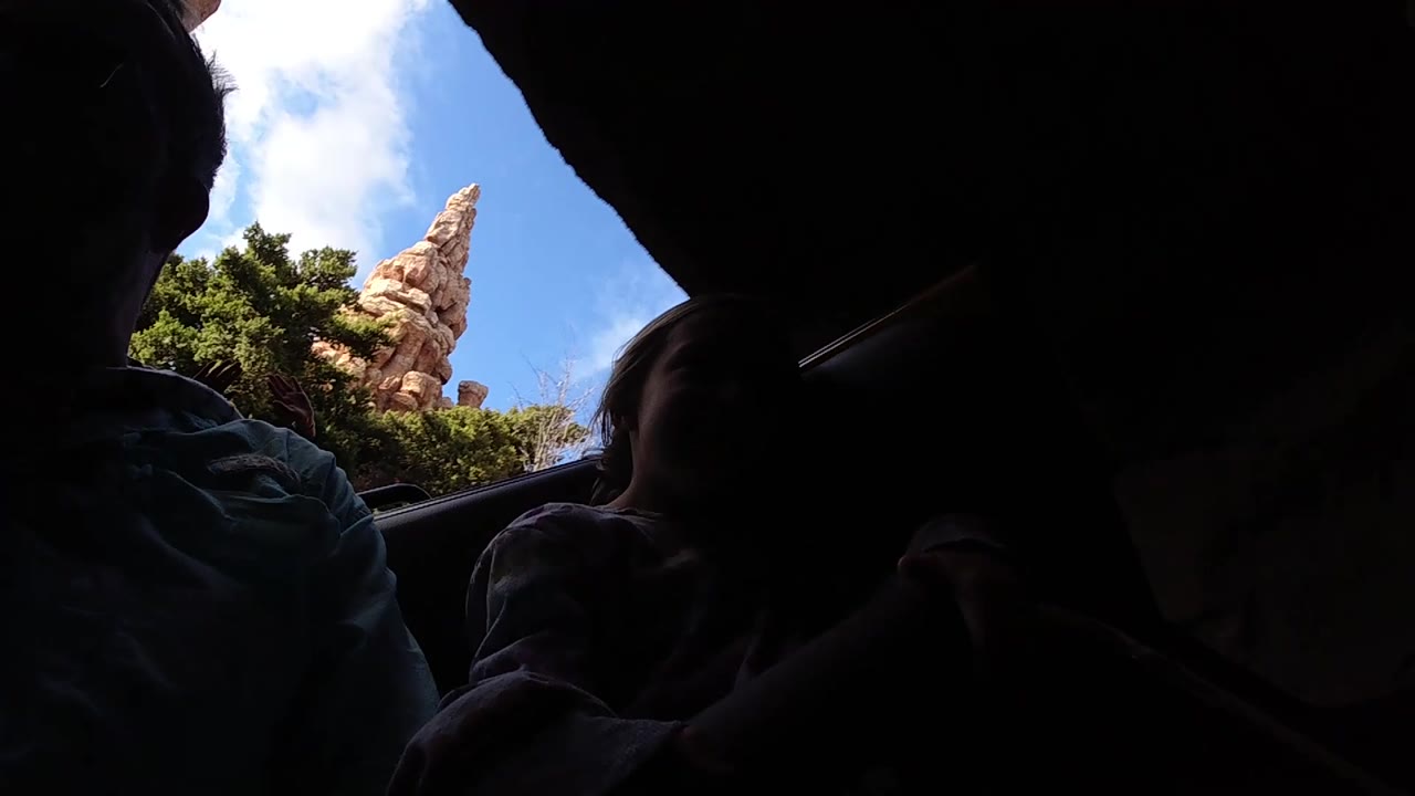 At Disneyland California, the camera sits low among the riders, with most faces in shadow and a sliver of blue sky and rockwork at the top right.