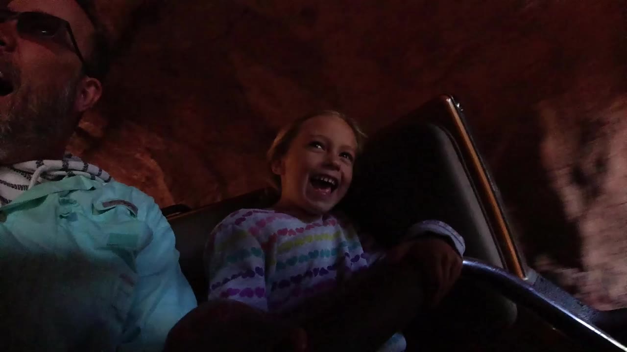 At Disneyland California, a young girl smiles beside a partially cropped adult man inside a ride vehicle under dark red rock-like walls.