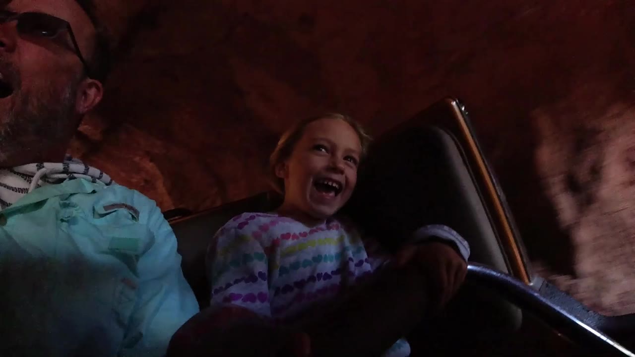 At Disneyland California, the girl laughs while seated beside an adult man, with the ride vehicle seat and red rock-like tunnel wall filling the background.
