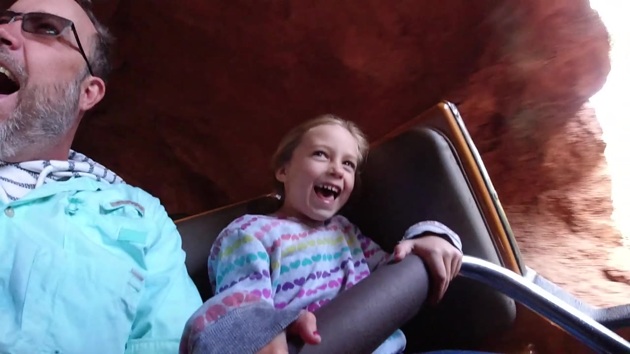 At Disneyland California, the adult man and young girl react with open mouths as bright daylight appears beyond the red rock-like ride structure.