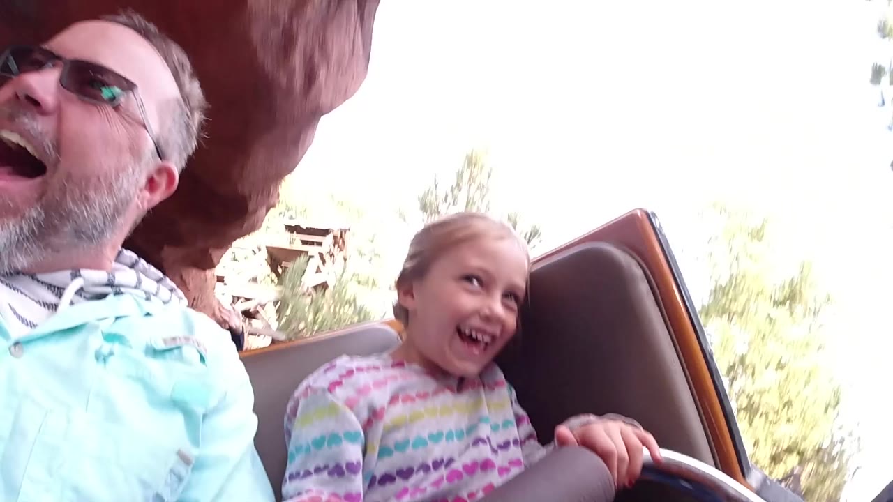 At Disneyland California, a father in sunglasses and a young daughter laugh in a brown ride car under reddish ride structure, with bright sky and trees behind them.