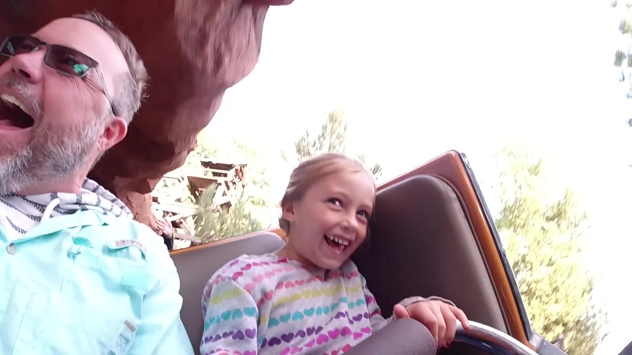 At Disneyland California, the father remains cropped at the left while the daughter smiles from the ride seat, with a leather-like side panel, pale sky, and trees in back.