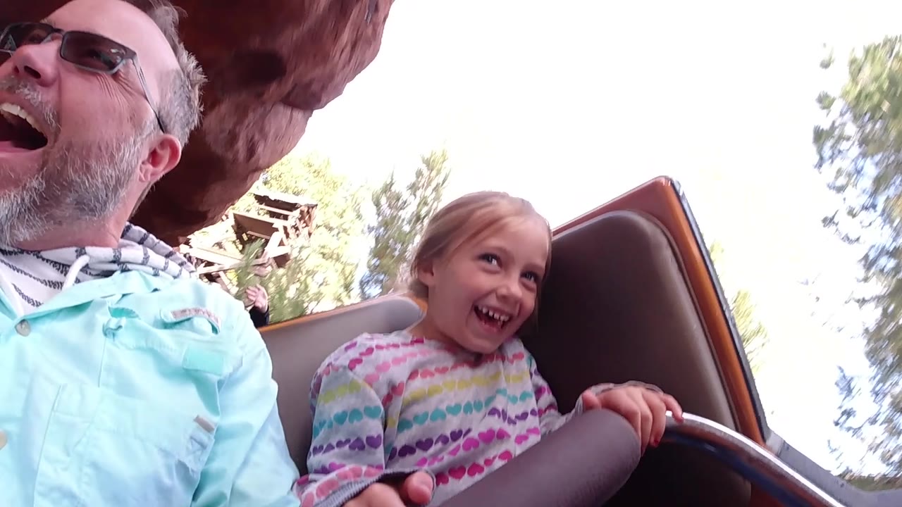 At Disneyland California, the father and daughter are more centered in the ride car as both laugh, with the daughter's hands near the front rail and trees visible to the right.