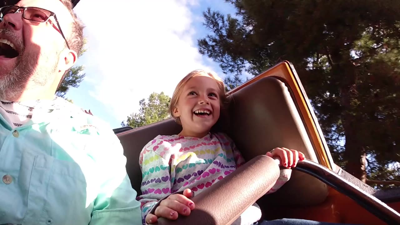 A father in sunglasses and a smiling young girl sit close together on a children's roller coaster at Disneyland California, with the safety bar and trees visible.