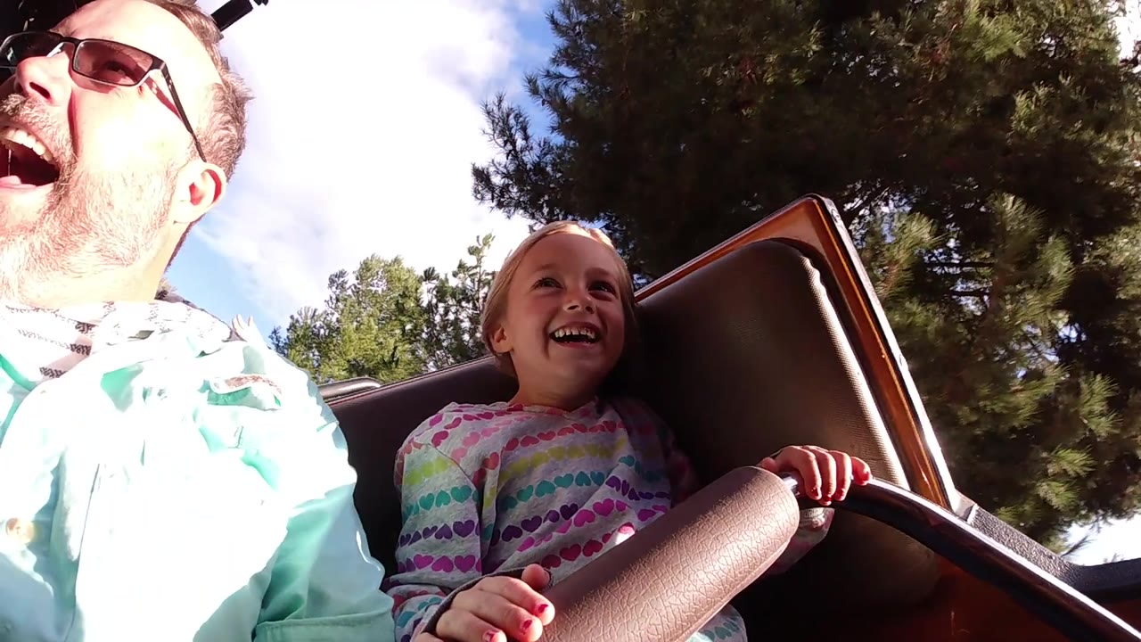 The father laughs with his mouth open while the young girl smiles beside him on the Disneyland California roller coaster, backed by trees and bright sky.
