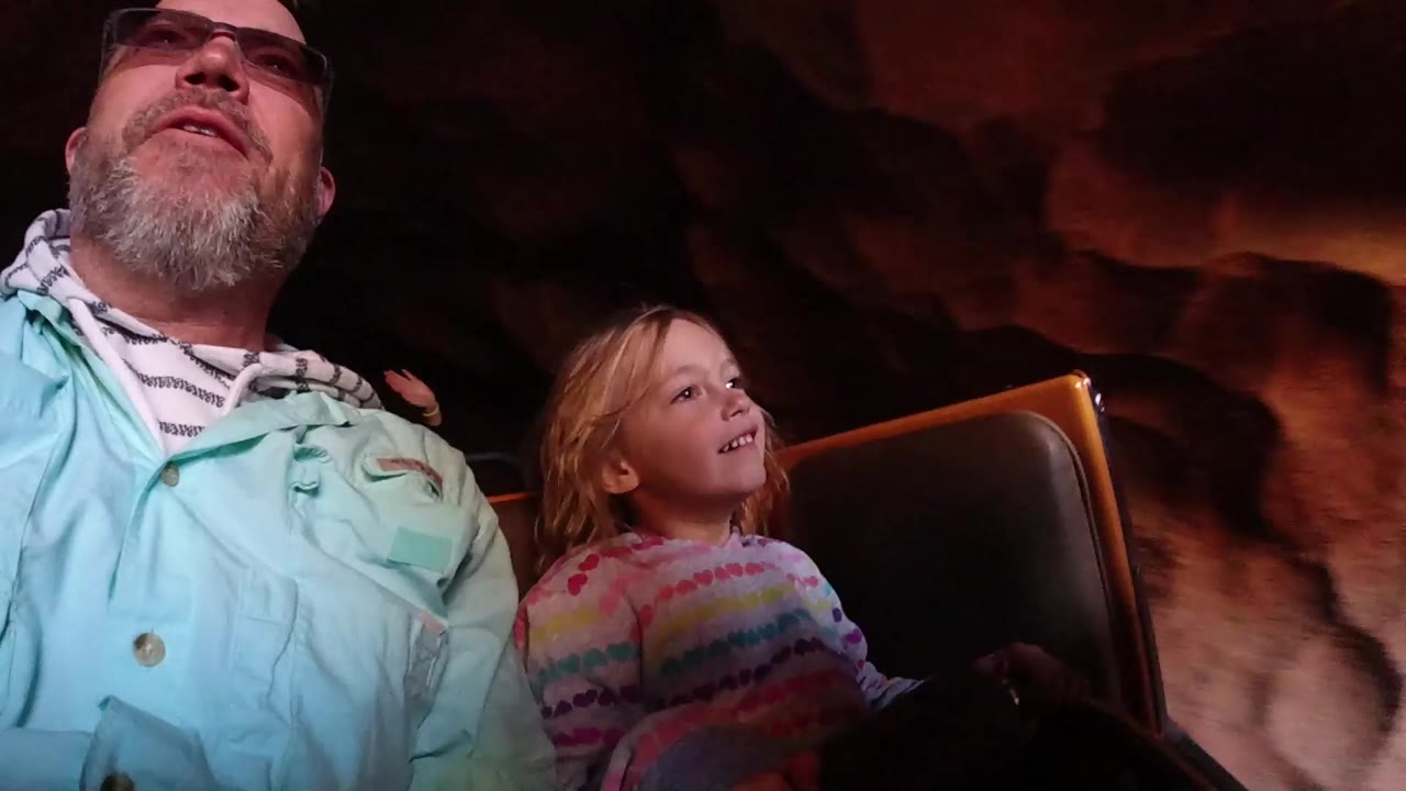 At Disneyland California, a father in glasses and a young child sit in a roller coaster car as dark reddish rock walls fill the background.