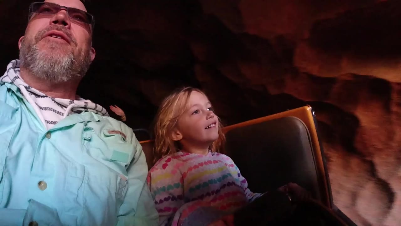At Disneyland California, the child smiles and looks up from the roller coaster car while the father sits beside them against shadowed red rock scenery.