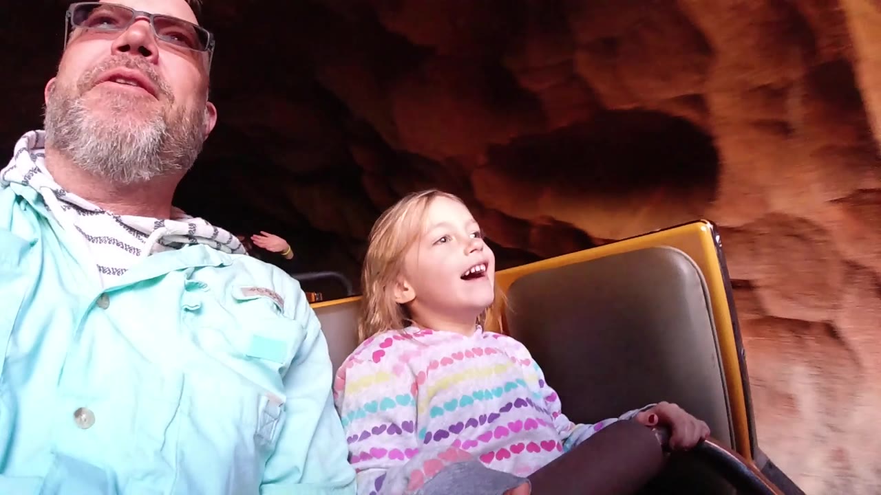 At Disneyland California, the roller coaster car passes into brighter light as the child smiles with one hand near the lap bar and the father looks ahead.