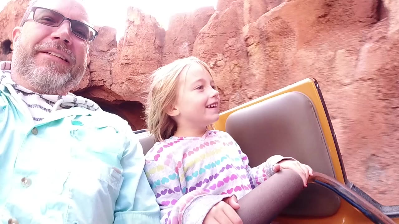 At Disneyland California, a father in glasses and a young daughter sit close together in an open ride car as red rock walls fill the background.