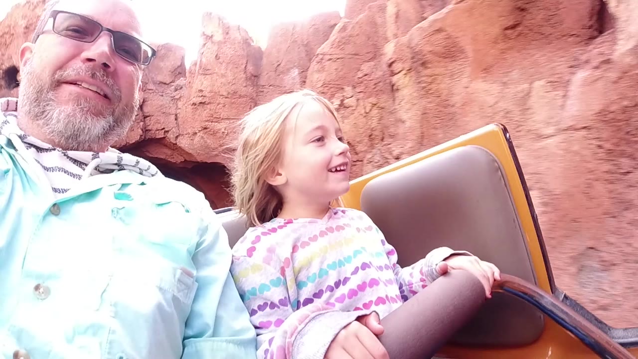 Father and smiling daughter ride past red rock scenery in an open car.