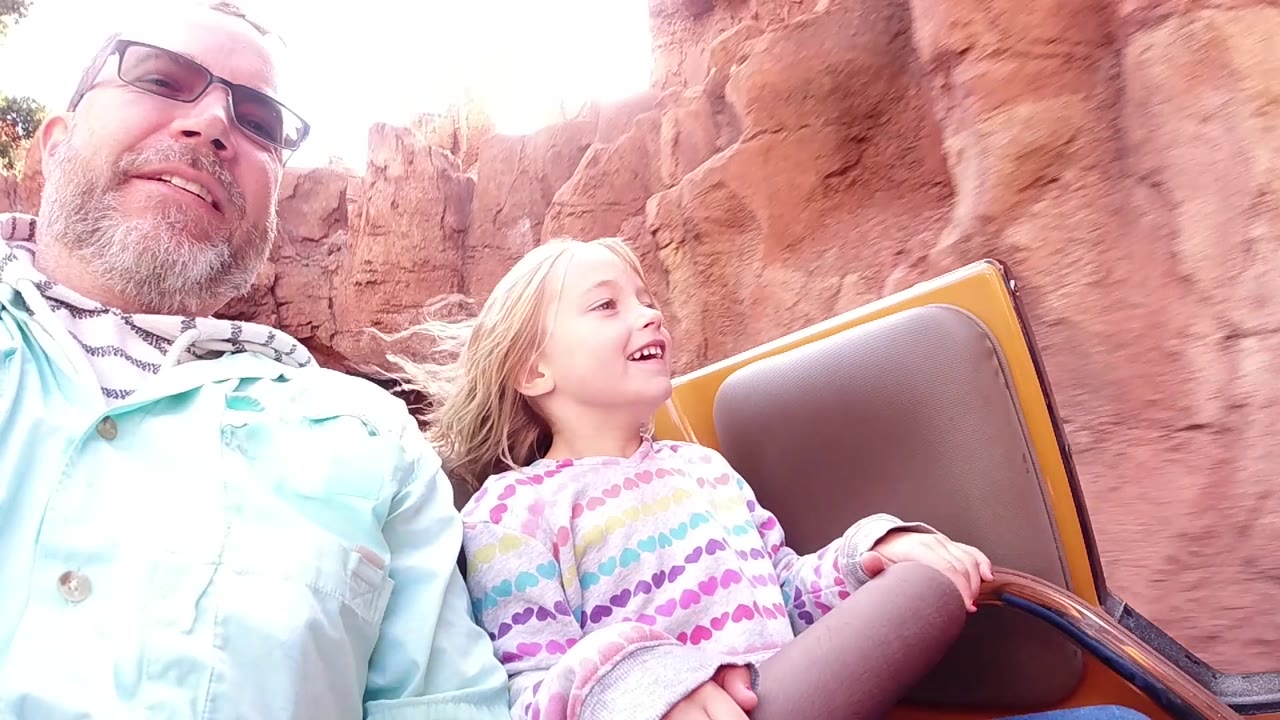 At Disneyland California, the father and daughter look ahead from the ride car, with the child's hair lifted by wind and red rock walls close behind.