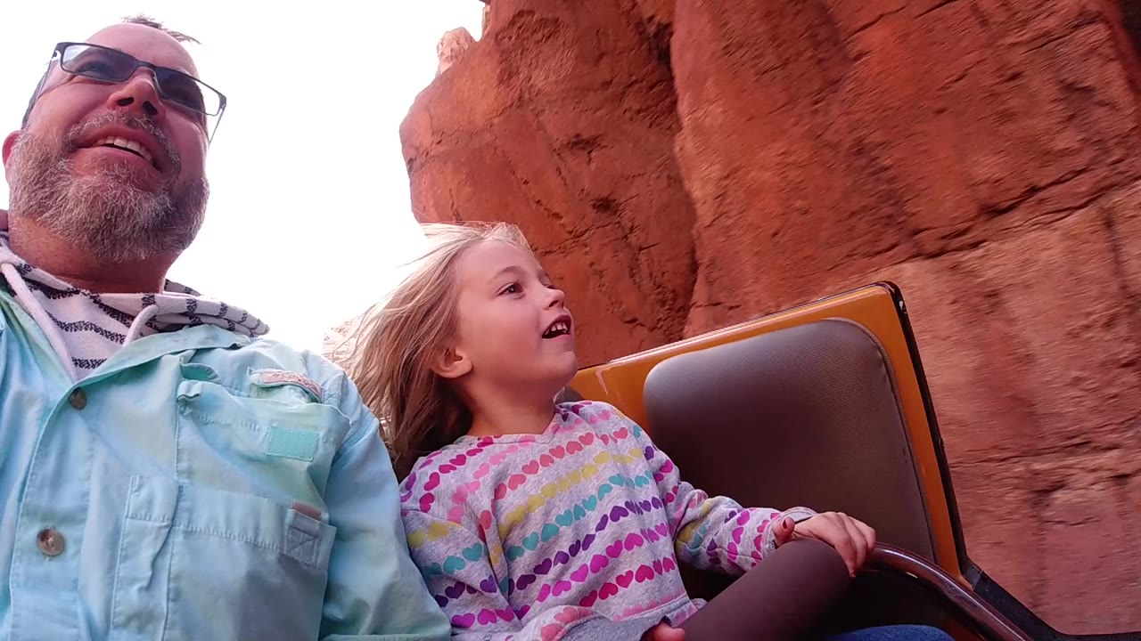 At Disneyland California, a man in sunglasses and a blonde child sit in a roller coaster car beside a close red rock wall.
