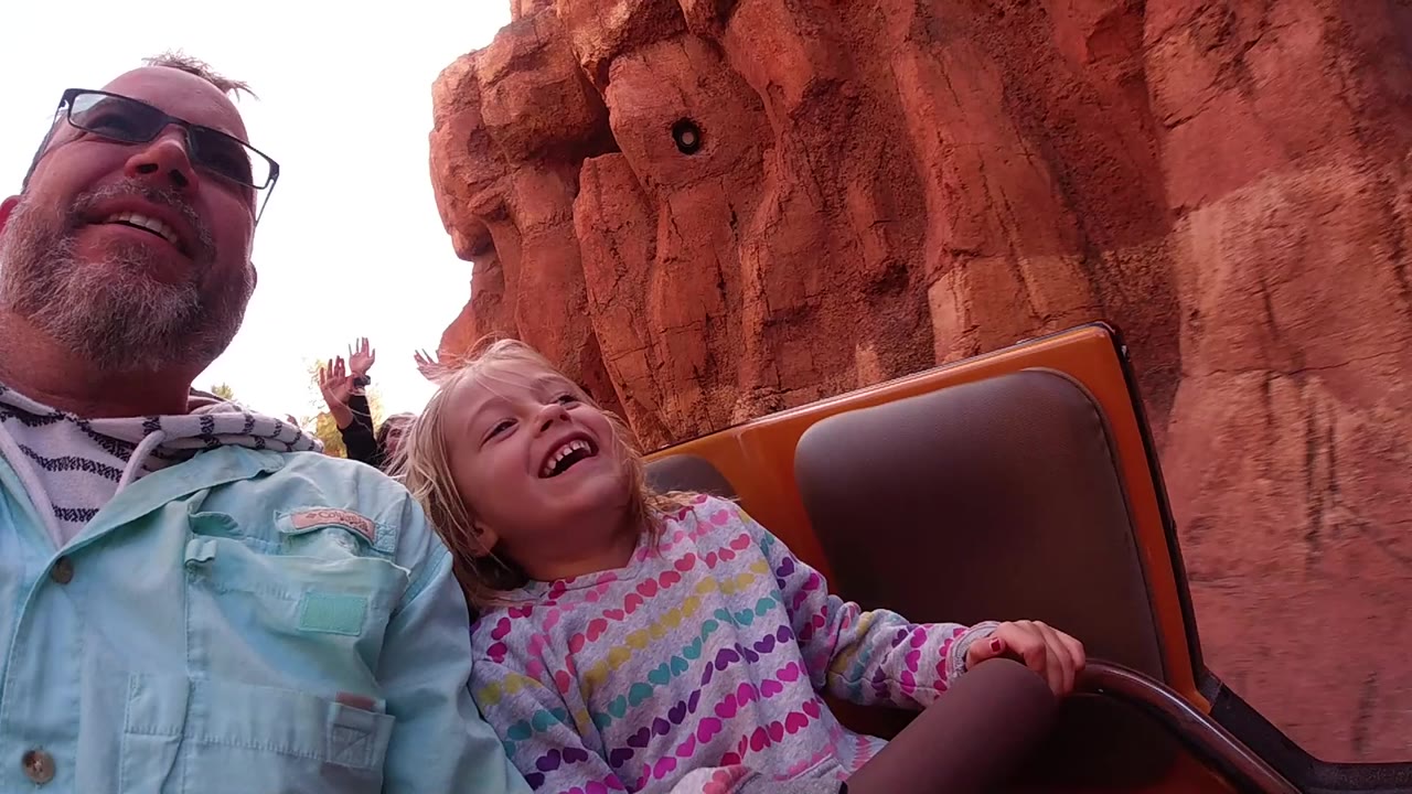 Man and child smiling in a theme park ride car with rockwork behind them.