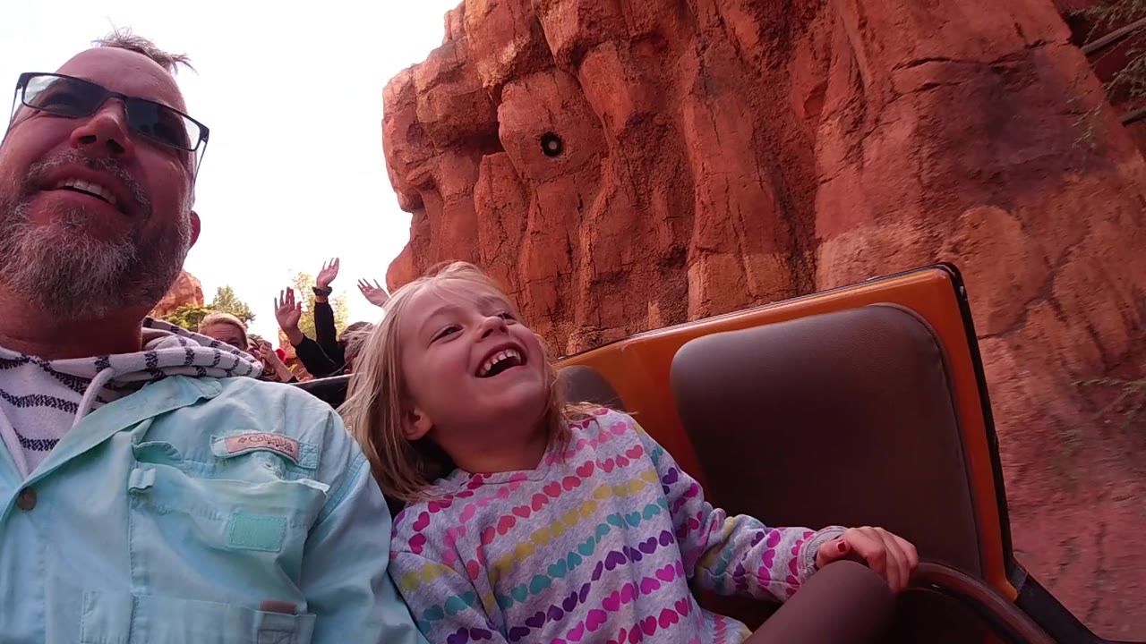 At Disneyland California, the child laughs with her head turned slightly upward as the man sits beside her and raised hands from other riders appear in the background.