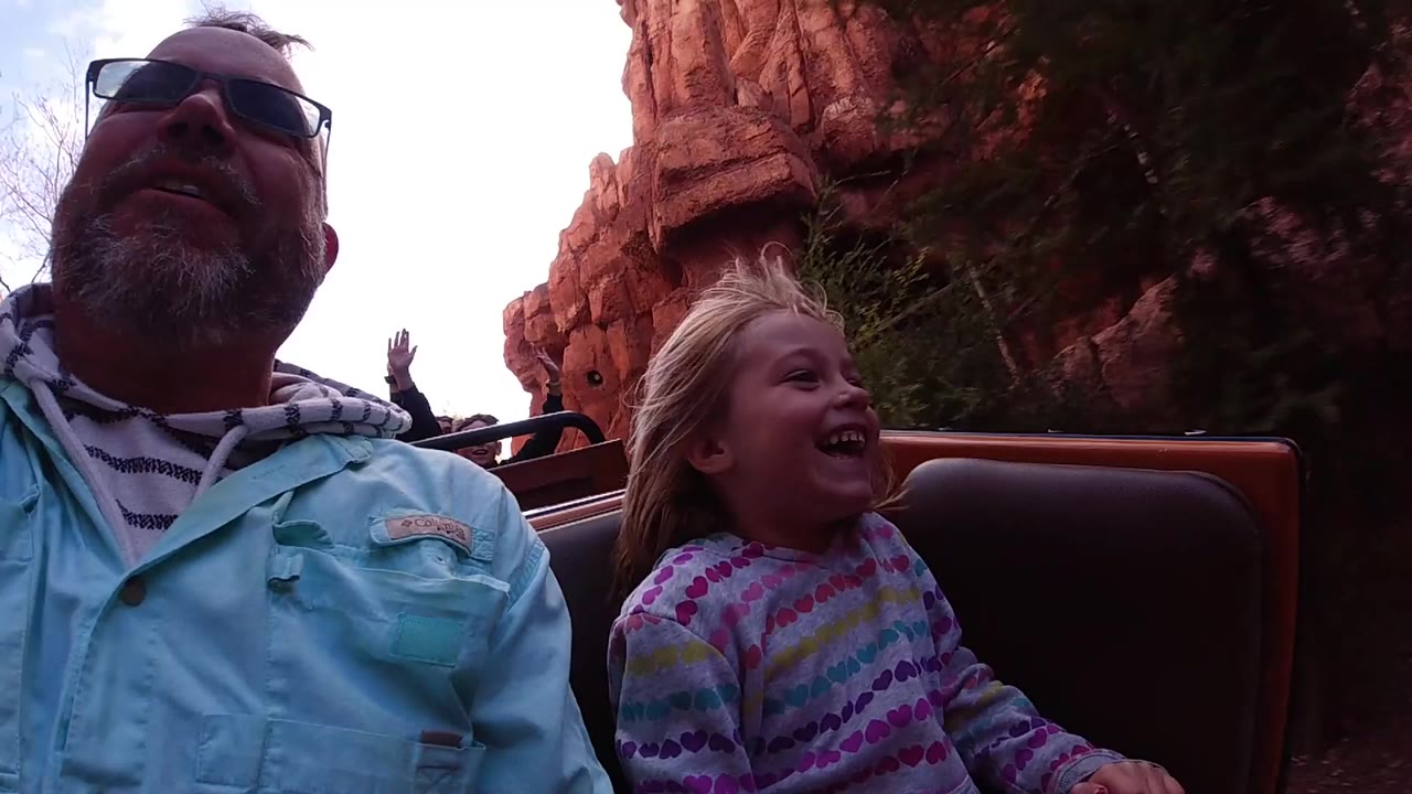 At Disneyland California, the young girl smiles beside the adult rider while red rock formations and a raised hand from another guest appear in the background.