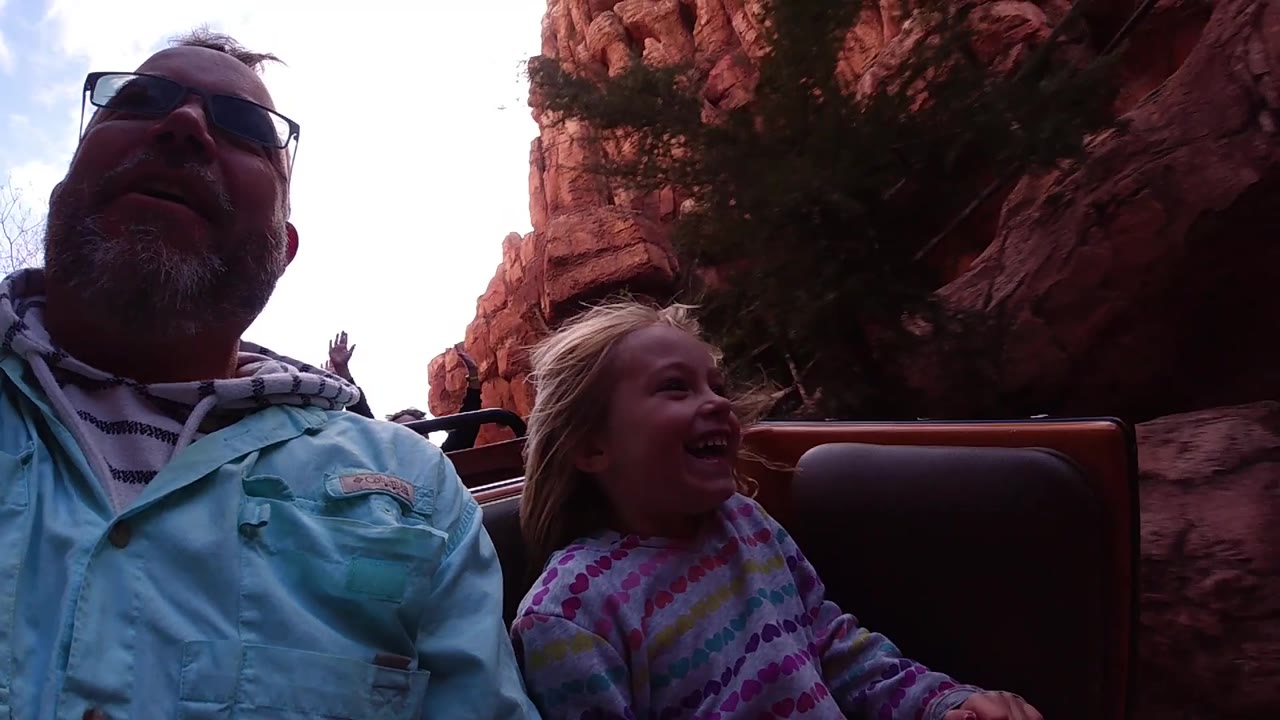 At Disneyland California, the adult and child remain in the foreground as the attraction vehicle moves past darker red rock walls and a seat back fills the center.