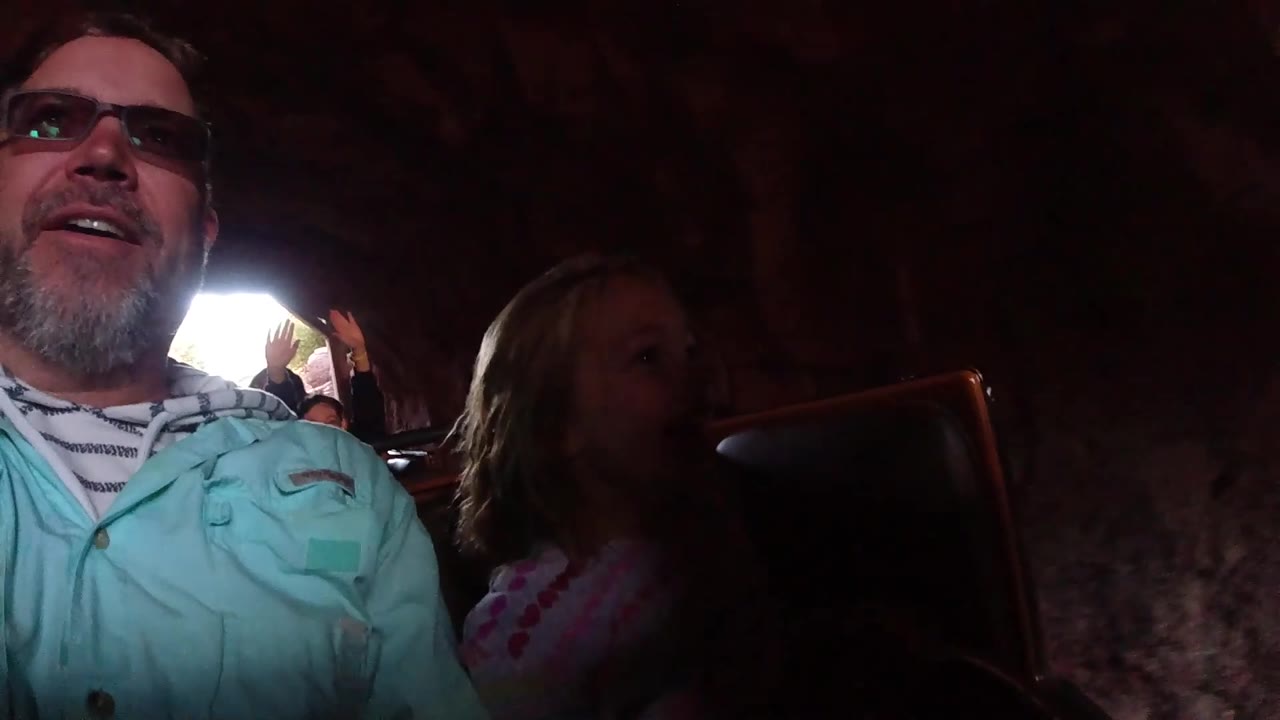 A father in glasses and a young blonde girl sit side by side in a Disneyland California coaster car, with reddish rock walls and bright daylight breaking into the frame.