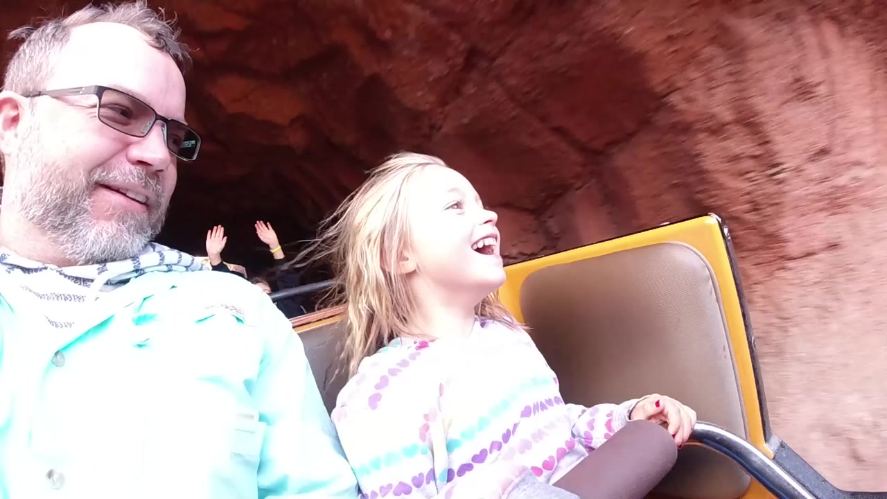 At Disneyland California, a blonde child smiles beside an older man in glasses as their ride vehicle passes reddish rockwork and seated riders behind them.
