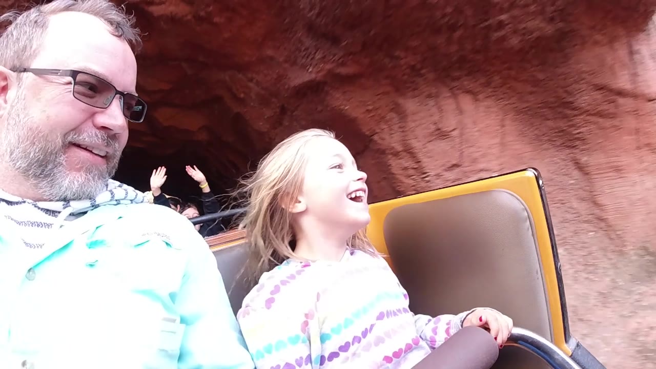 At Disneyland California, the child smiles toward the right edge of frame while an older man sits to her left and red rockwork curves behind the ride vehicle.