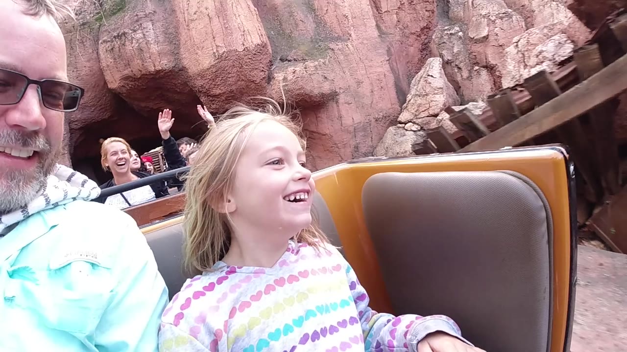 At 0:01 in the Disneyland California clip, the girl's face turns toward the ride path while the adult man remains partly in frame and the brown seat back fills the foreground.