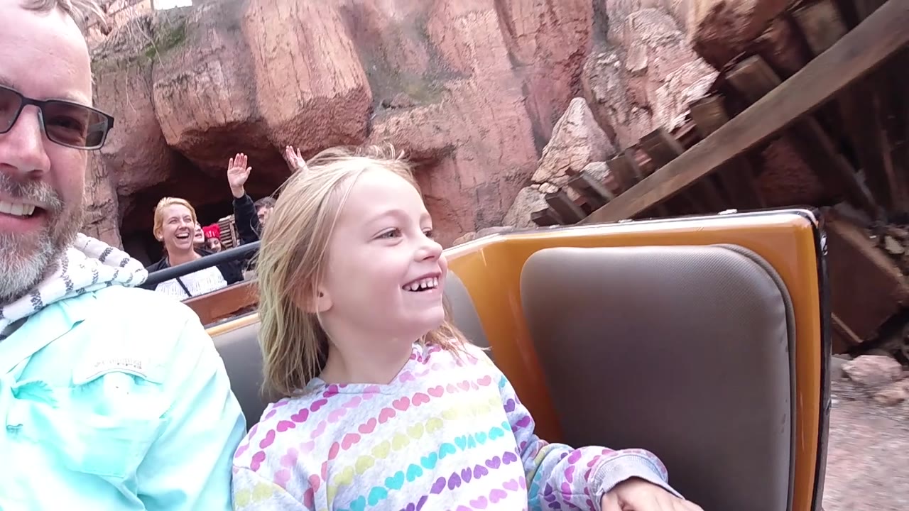 At Disneyland California, the young girl continues smiling in the coaster car as the adult man appears closer to center and reddish rock formations and dark track supports sit behind them.