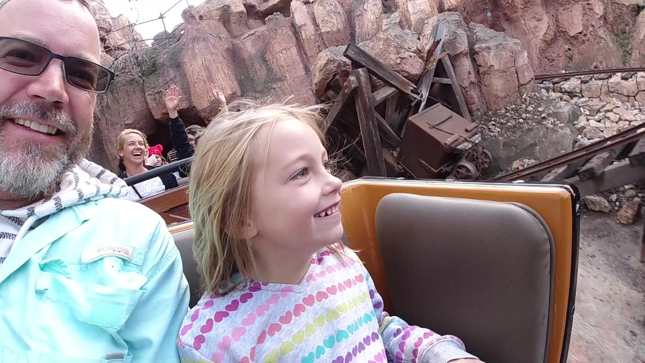 At Disneyland California, a father in glasses and a young blonde daughter smile from a brown ride car with rocky themed scenery and timber props behind them.