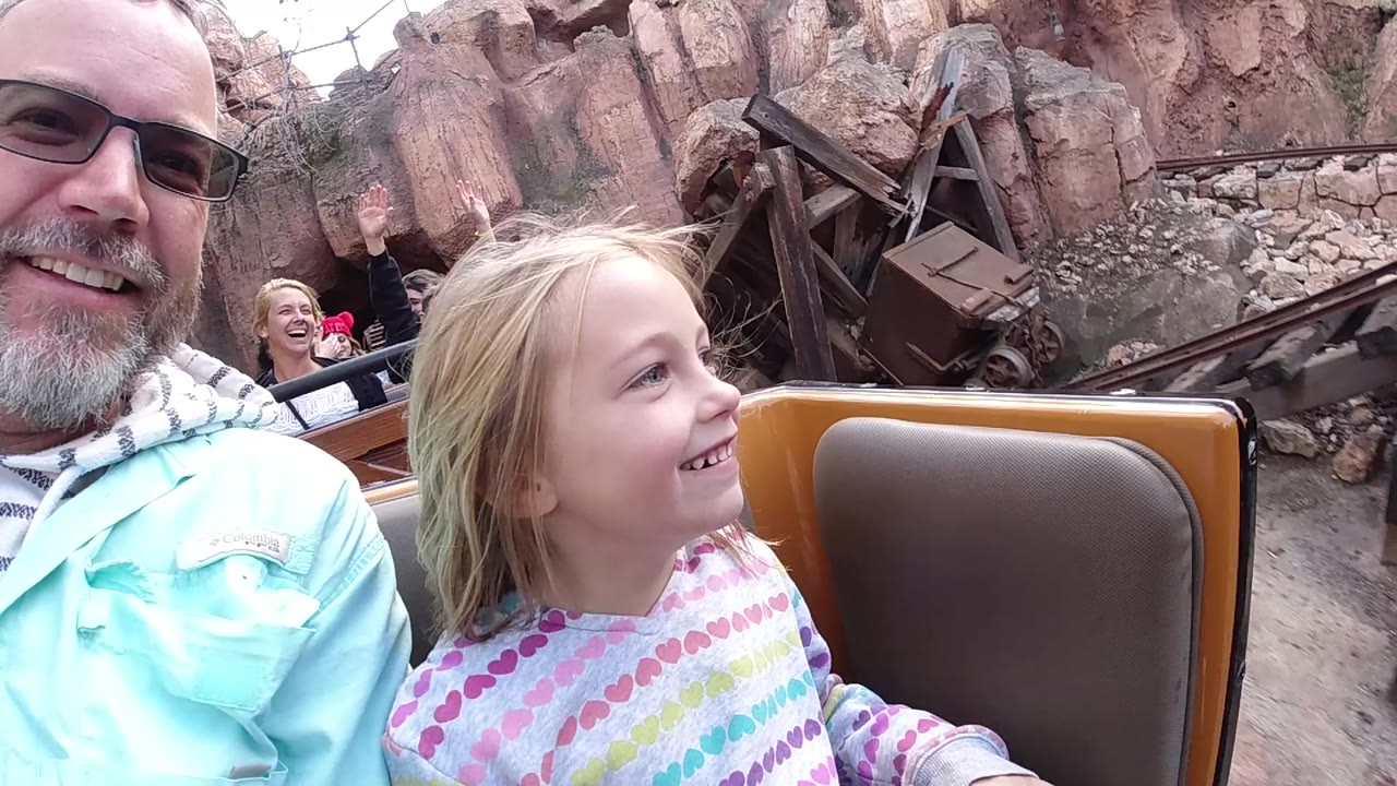 The father and daughter remain close to camera in the Disneyland California ride car while a woman behind them raises a hand near rockwork and mining-themed set pieces.