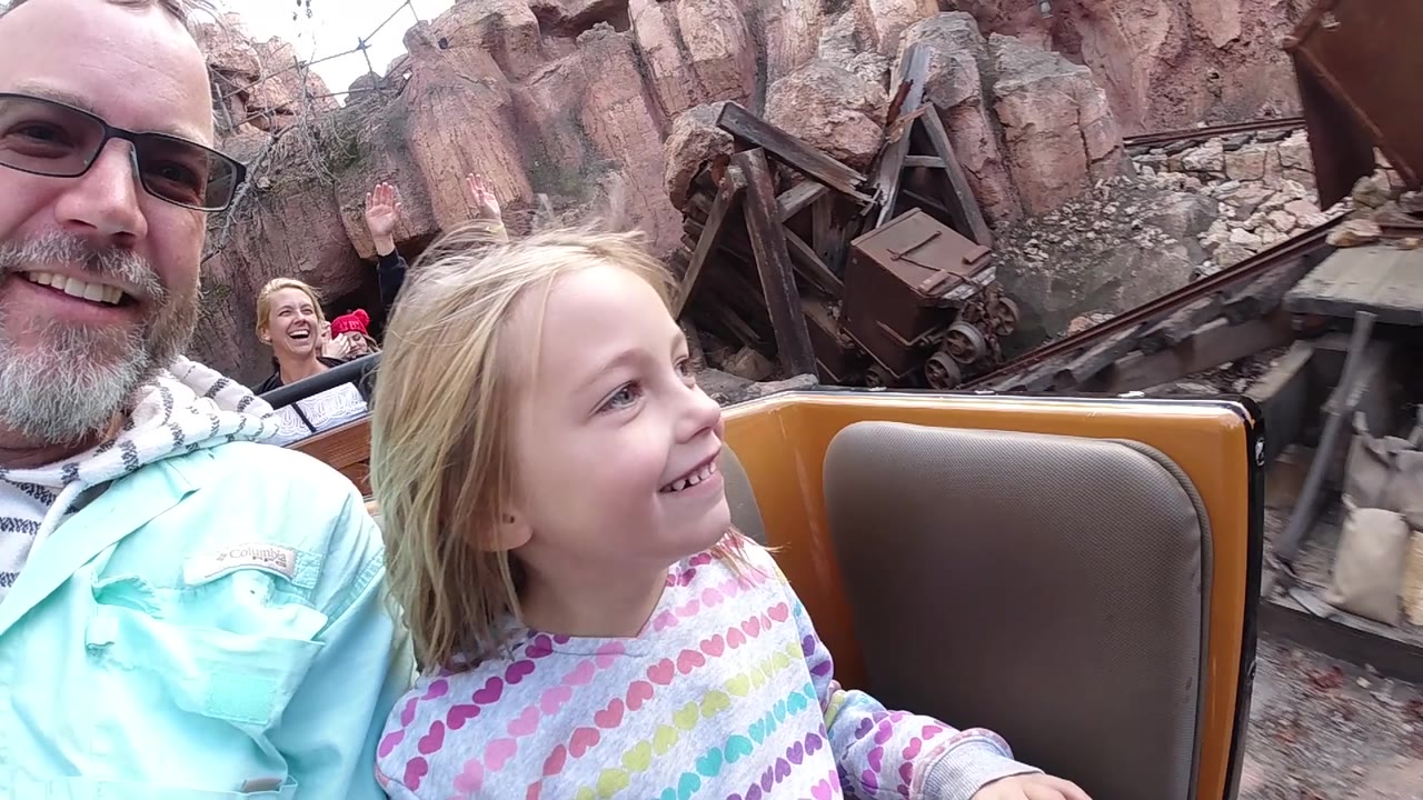 A young girl in a striped heart-pattern top looks forward from the Disneyland California ride car as her father smiles at frame left and a mine-cart prop fills the rocky background.