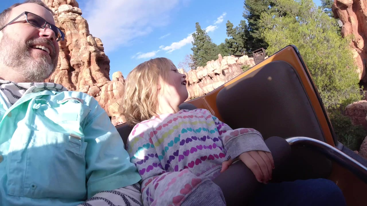 At Disneyland California, a bearded man in sunglasses and a young blonde child sit low in a roller coaster car with orange rockwork and blue sky behind them.