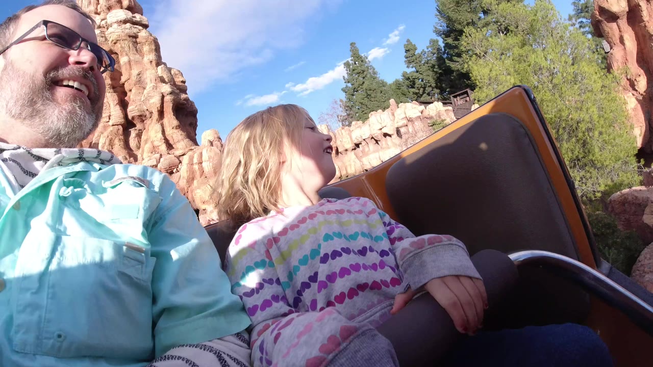 At Disneyland California, the man laughs near the left edge while the child turns toward the coaster car side panel, with rock formations and trees passing in the background.