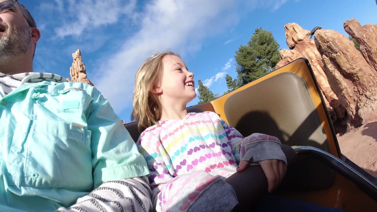 At Disneyland California, the child smiles toward the right side of the frame while the man is partly cropped, with tan-orange rock spires, trees, and blue sky visible.