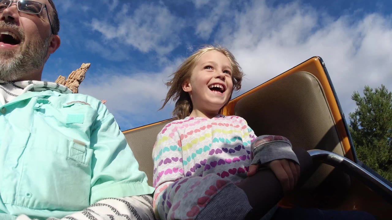 Young child laughs in a roller coaster seat under a cloudy blue sky.