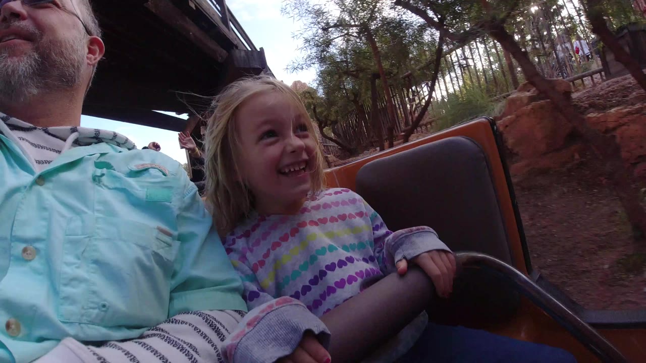 At Disneyland California, the girl remains seated next to the adult, smiling toward the ride path as trees, railings, and reddish landscaping pass in the background.