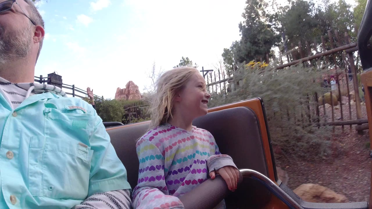At Disneyland California, the girl laughs while holding the seat rail, with the adult cropped at left and shrubs, fencing, trees, and pale sky visible behind the ride car.