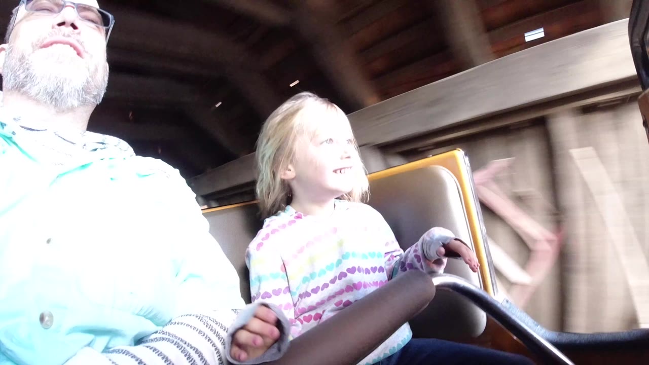 At Disneyland California, the father and daughter remain seated in the coaster car as bright light washes the foreground and a wooden or metal ride structure passes behind them.