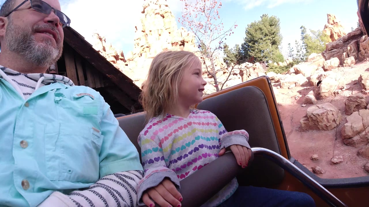 At Disneyland California, the coaster car is outdoors with the father and daughter in the foreground, reddish themed rocks and trees behind them, and blue sky above.
