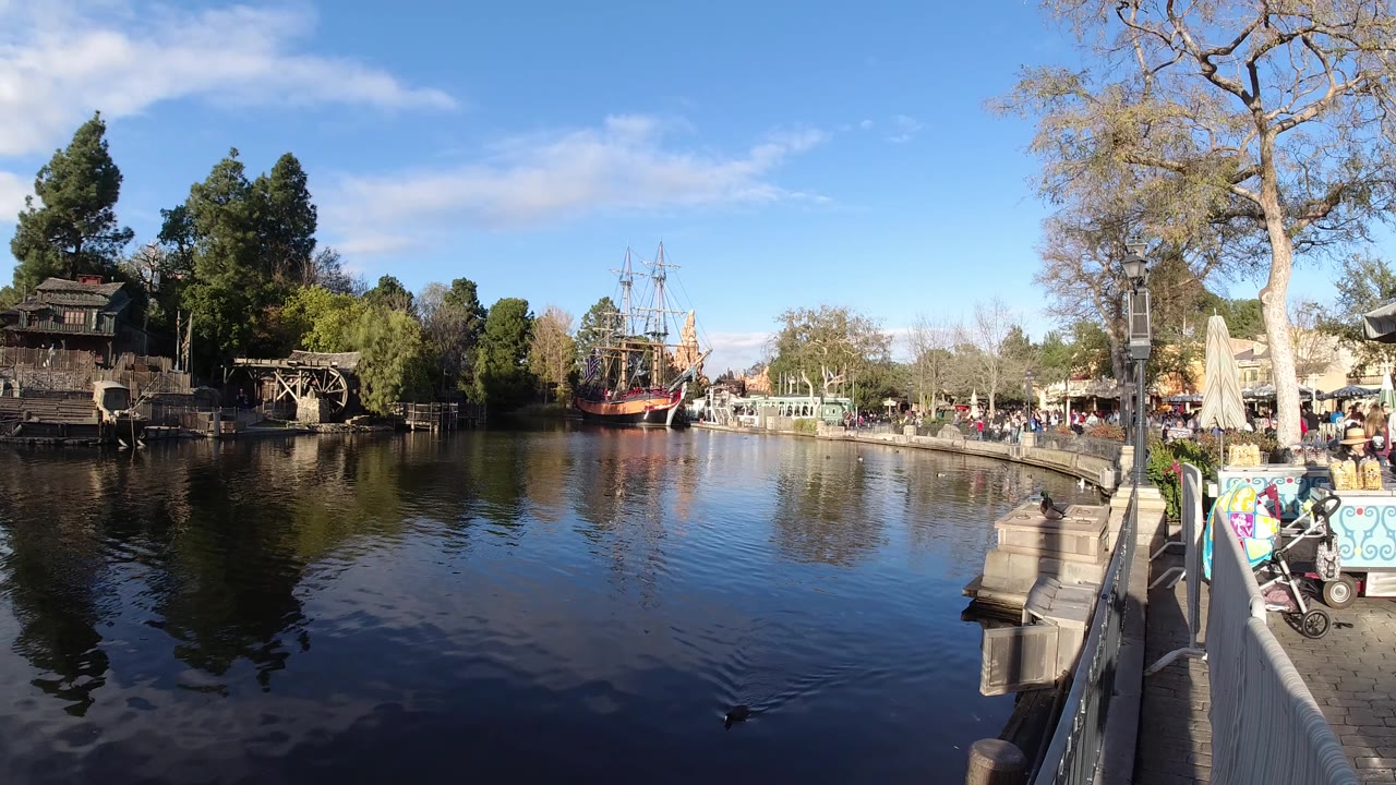 At 0:01 in Disneyland California, the lake surface shows small ripples below trees, a foreground railing, and a red sailing ship set back near the opposite shore.