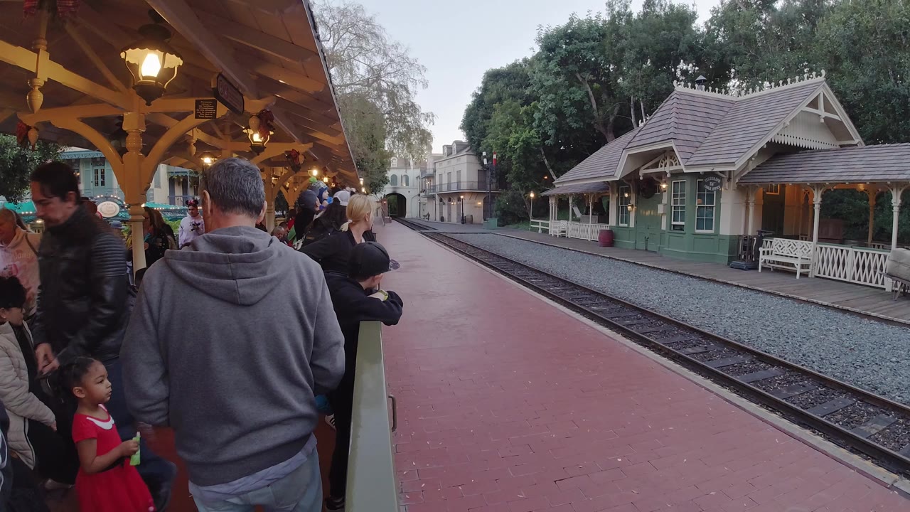 At Disneyland California, adults and children stand under a wooden station canopy beside red pavement, train tracks, a green station building, and trees.