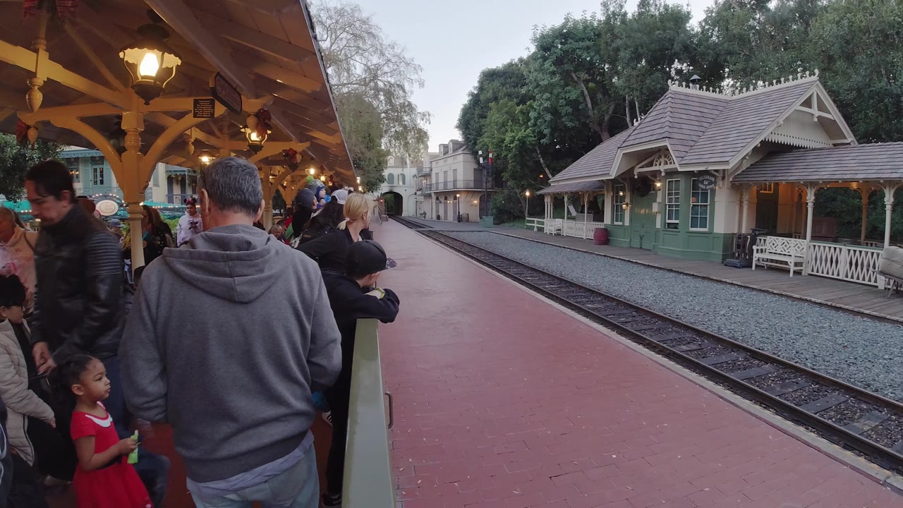 People stand on a red platform beside train tracks and a green station building.