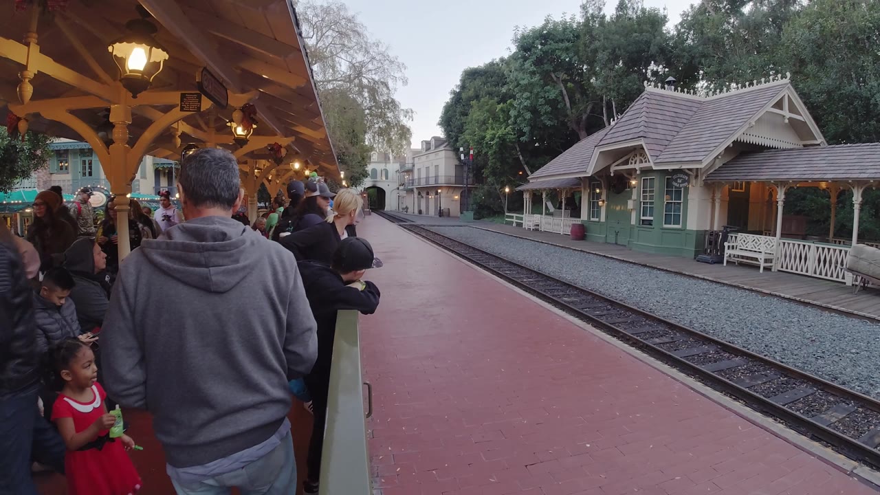 At Disneyland California, a man in a gray hoodie faces the tracks while families gather under warm station lights near the green station structure.