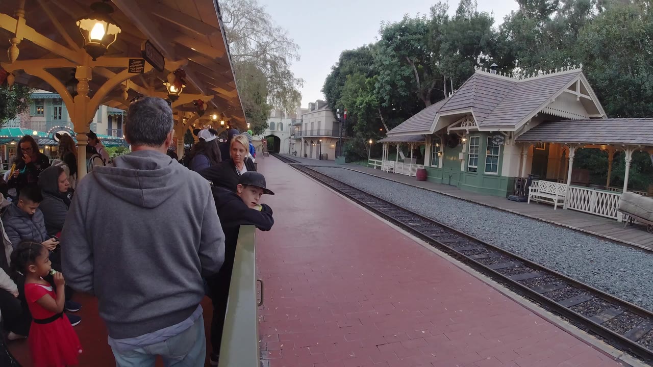 People line the platform railing across from a small station building.
